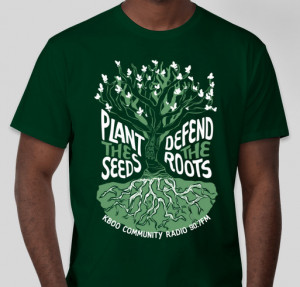 Green t-shirt worn by model. Shirt has an illustration of a tree with text surrounding the tree reading "Plant the Seeds, Defend the Roots"