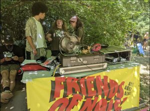 THree young people chat behind a music production console underneath trees on a sunny day