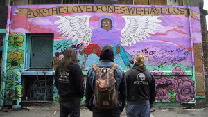 Three people stand in the foreground, backs to camera, looking at a wall mural entitled "For the Loved Ones We Have Lost" that features an angel holding someone in their arms.