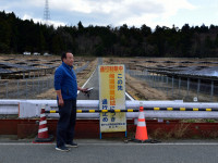 Takeshi Yamakawa of Tokyo Shimbun stands next to a sign on Route 253 near Namie that says, “Exclusion zone: do not enter.” A mega-solar farm built on abandoned farms is on the other side of the highway fence. (Photo courtesy of U.G. Kaneko) Takeshi Yamakawa of Tokyo Shimbun stands next to a sign on Route 253 near Namie that says, “Exclusion zone: do not enter.” A mega-solar farm built on abandoned farms is on the other side of the highway fence. (Photo courtesy of U.G. Kaneko)